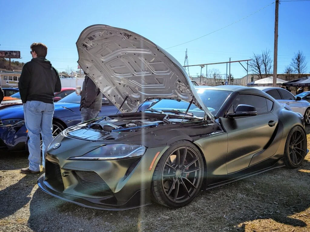 Matte gray Toyota Supra with open hood at outdoor car event with people and other vehicles in background