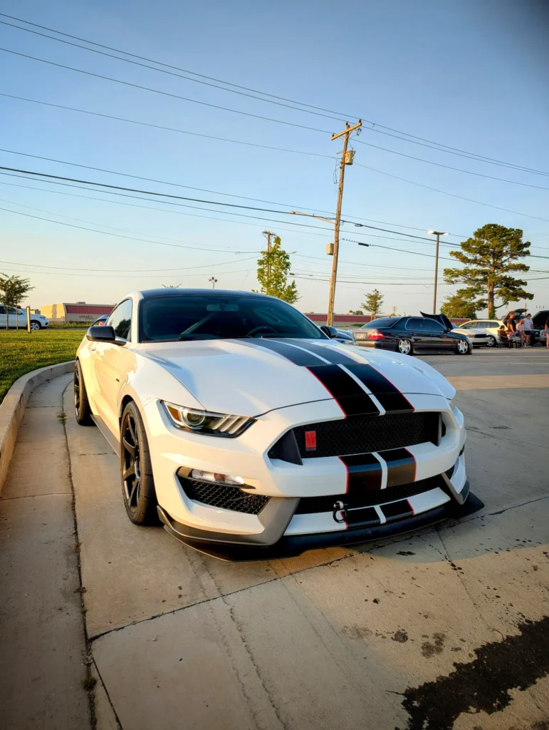 White Ford Mustang Shelby GT350 with black and red racing stripes parked in a lot during late afternoon at a car event