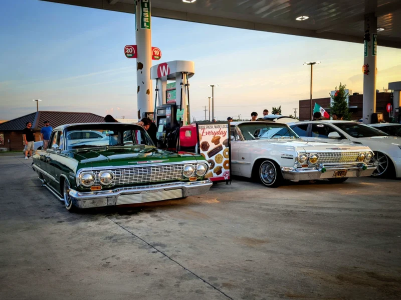 Green and white Chevrolet Impalas parked side by side at a gas station with people and a Mexican flag nearby
