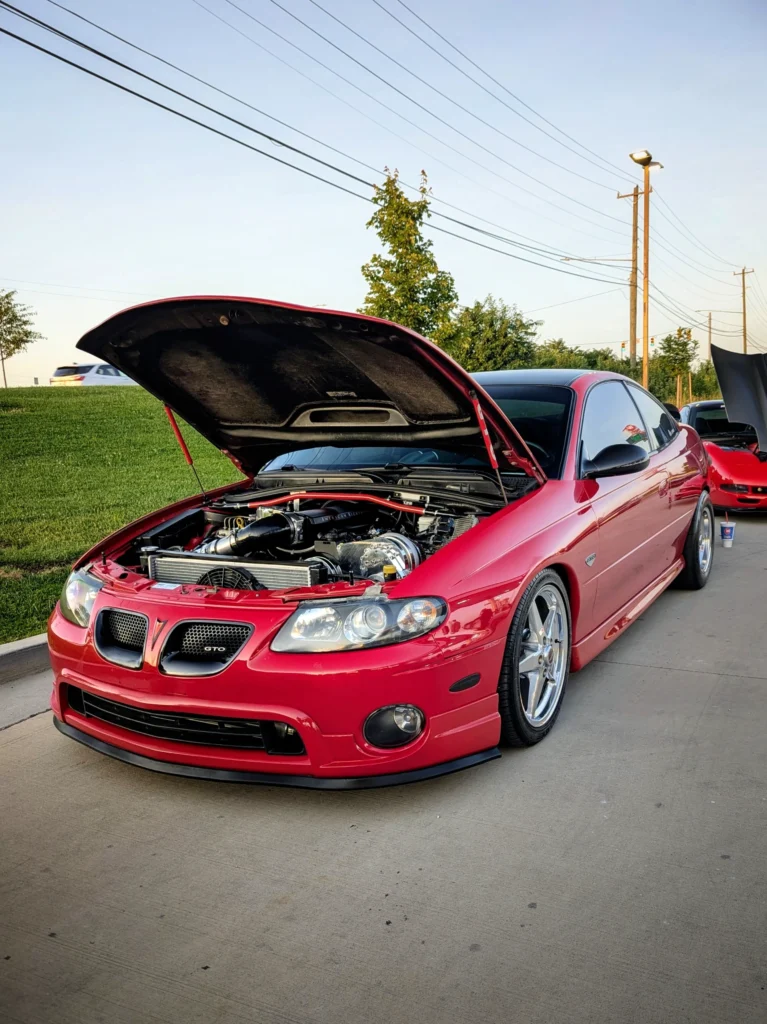 Red Pontiac GTO with open hood showing engine, parked on concrete near grass and trees