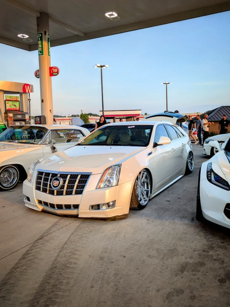 White Cadillac sedan with lowered suspension parked at a gas station during a car meet with people and other vehicles in back