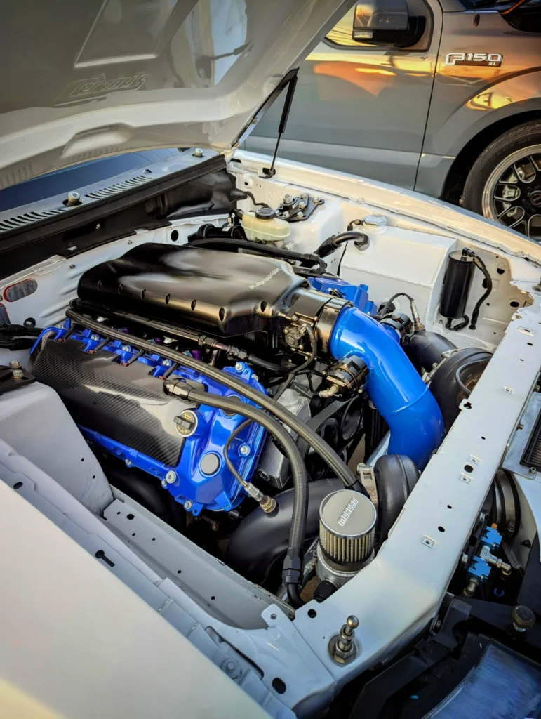 Clean engine bay with blue and black components in a white car, next to a gray Ford F-150 XL pickup at sunset