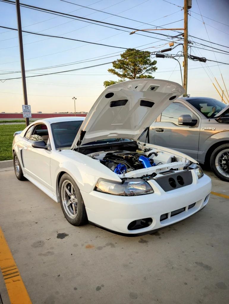 White Ford Mustang with hood raised showing engine, parked next to a silver Ford F-150 in a lot