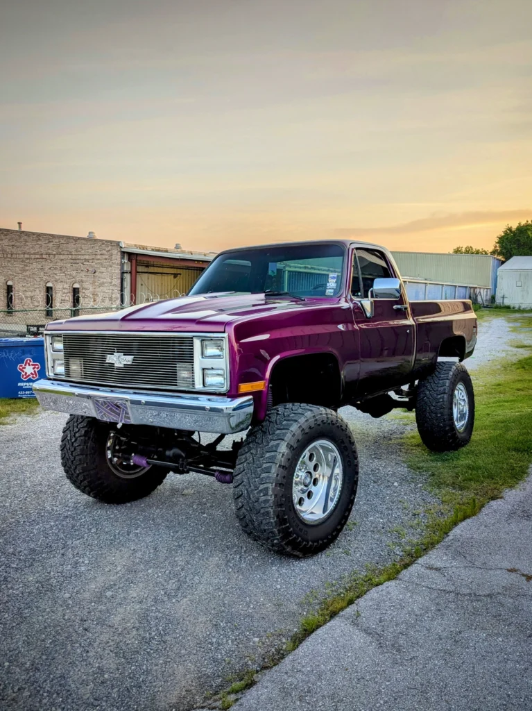 Purple Chevrolet K10 pickup truck with lift kit parked on gravel near industrial buildings at sunset