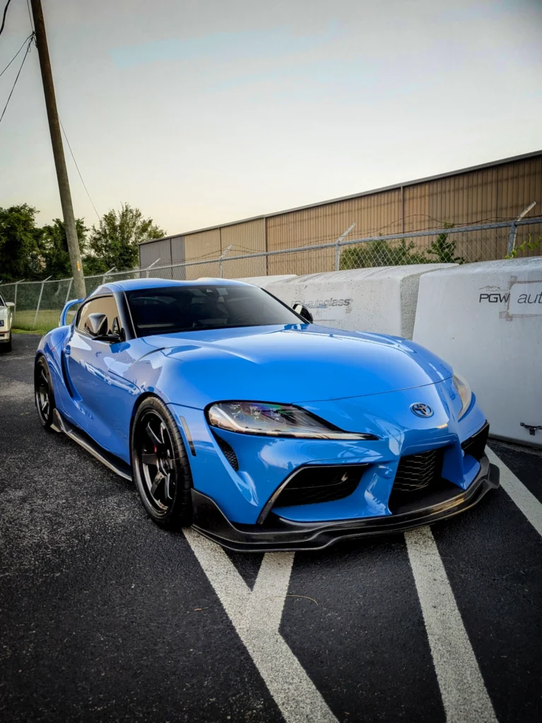 Blue Toyota Supra parked on asphalt with fencing and industrial building in background at dusk