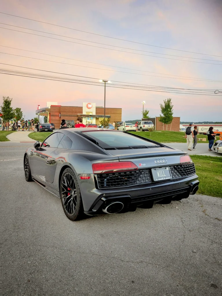 Gray Audi R8 parked at a car meet near a brick building with people and other vehicles in the background at dusk.
