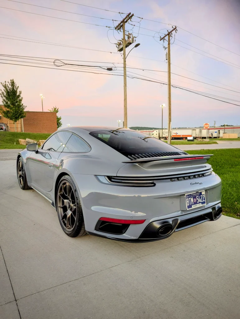 Silver Porsche 911 Turbo S parked on a concrete surface near a road and utility poles at dusk