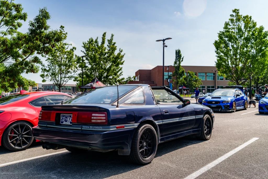 Black Toyota Supra parked among other cars at an outdoor car event with trees and buildings in the background