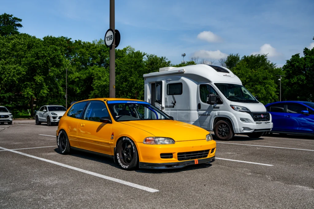 Yellow Honda Civic hatchback with black wheels parked in a lot with a white Ram Wingamm RV behind it and trees in the backgro