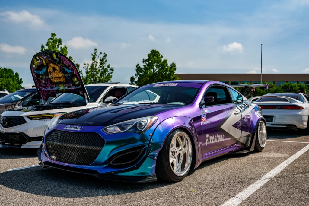 Purple and blue Hyundai Genesis with a wide body kit parked next to a white Acura Integra with its hood open at a car meet.
