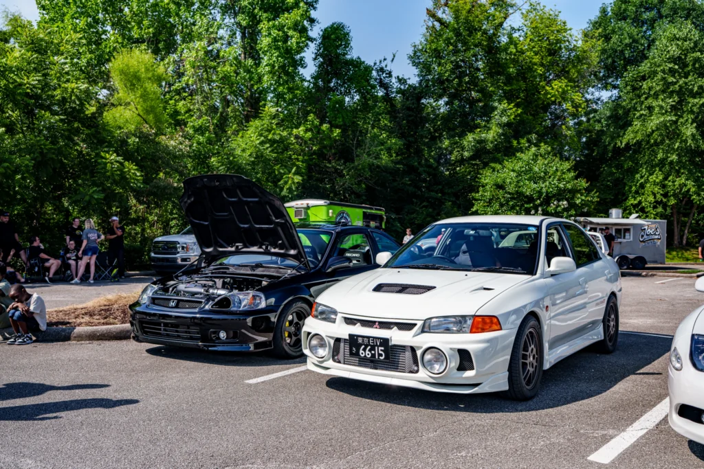 White Mitsubishi Lancer Evolution and black Honda Civic with hood open in a parking lot at a car meet with people and trees i
