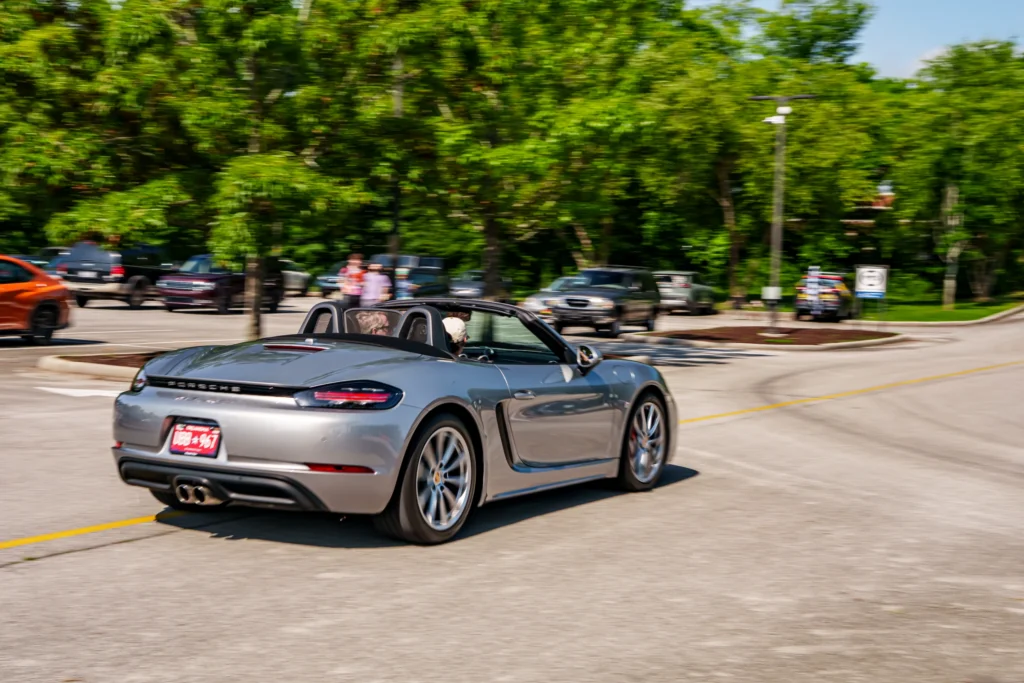 Silver Porsche 718 Boxster convertible driving down a street with two people inside and trees in the background