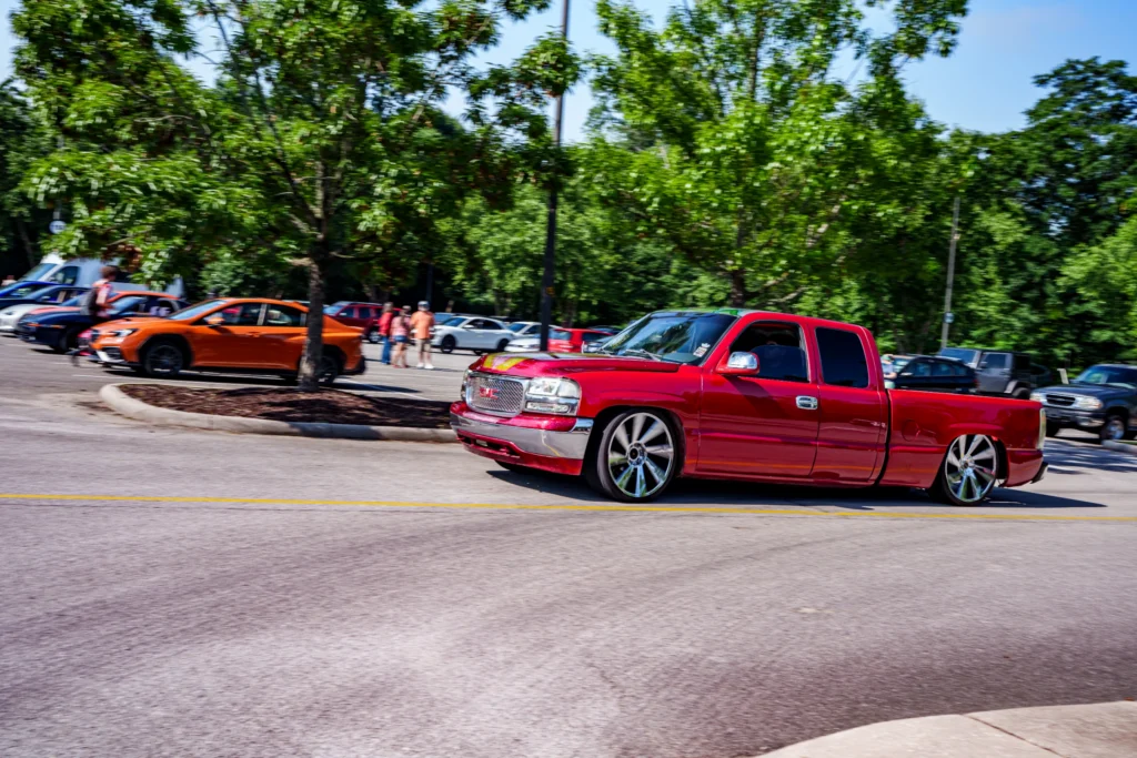 Red GMC pickup truck with large wheels driving on a road near parked cars and some people in trees and parking lot
