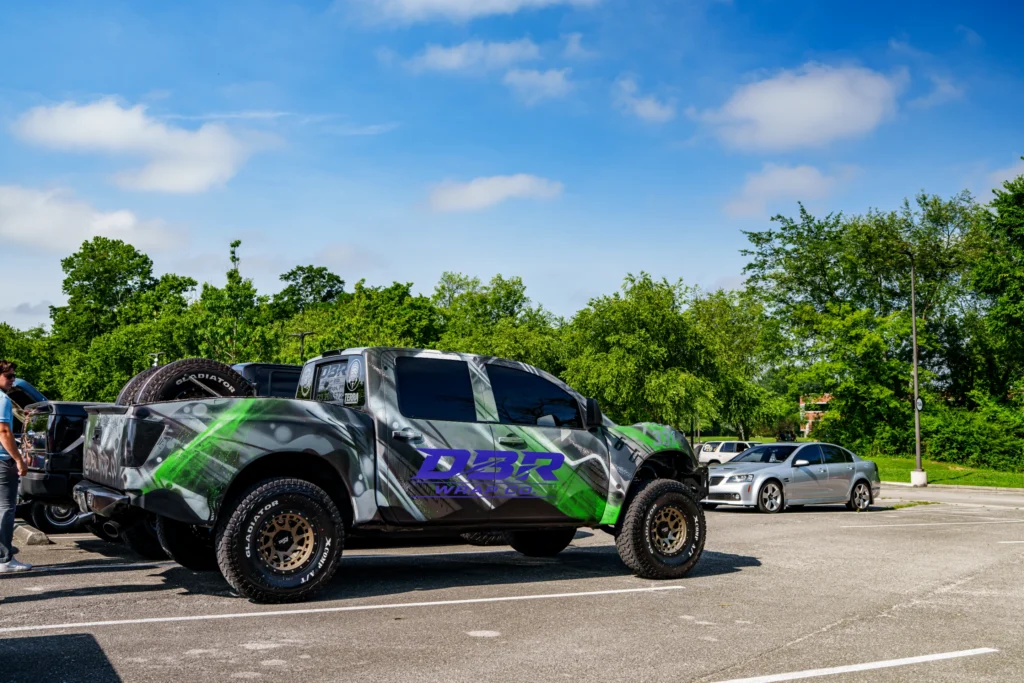 Gray camouflage wrap pickup truck with off-road tires and purple DBR Wrap Co. logo parked in lot with trees and blue sky in b