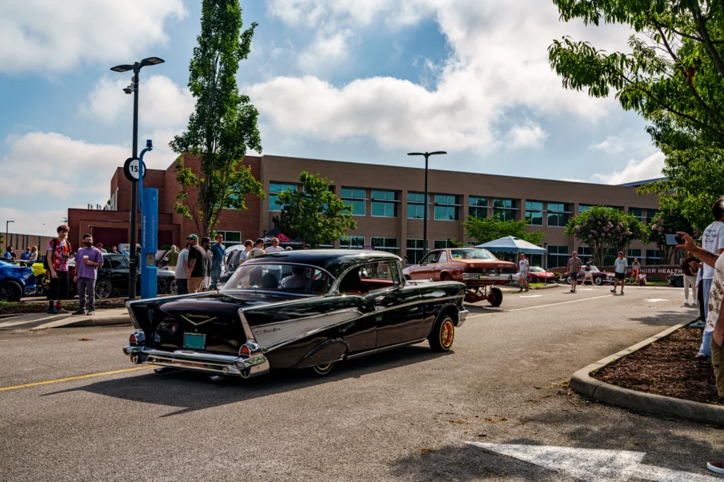 Black Chevrolet Bel Air driving on a street at a daytime car event with spectators and other vehicles around
