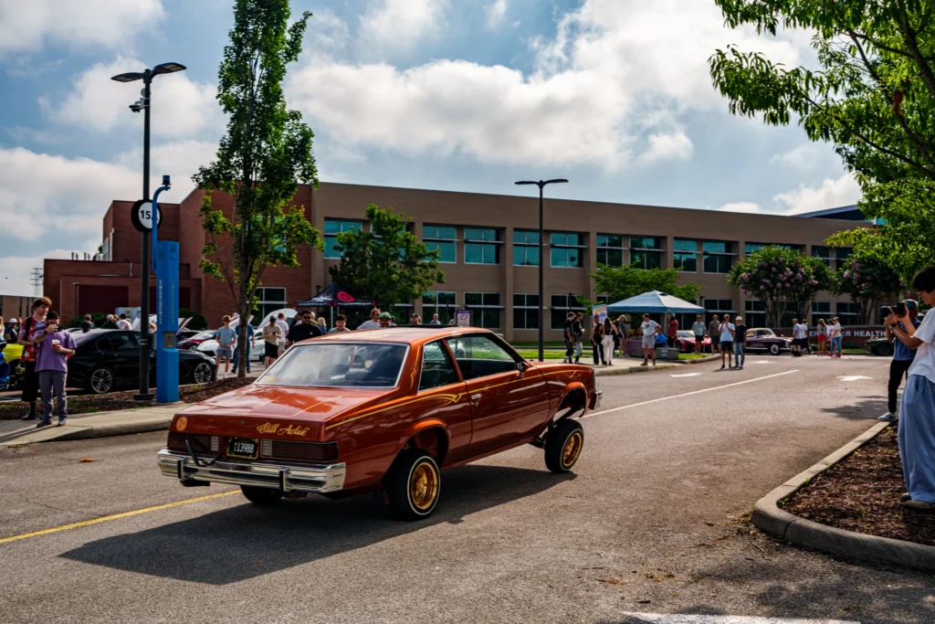Orange Buick Regal with raised front wheels driving down a street lined with spectators and tents under a partly cloudy sky.
