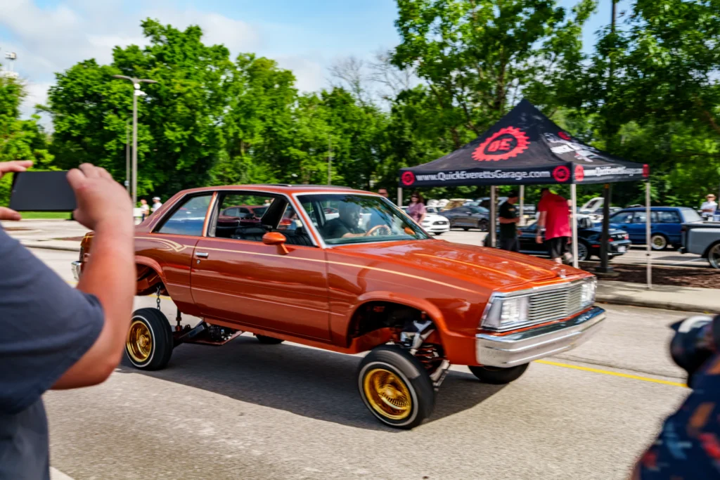 Orange raised Buick Regal with gold rims driving in a parking lot during a car event with people and tents in the background