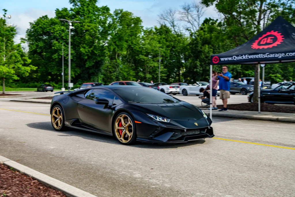 Black Lamborghini Huracán with gold wheels parked at an outdoor car event with people and tents in the background