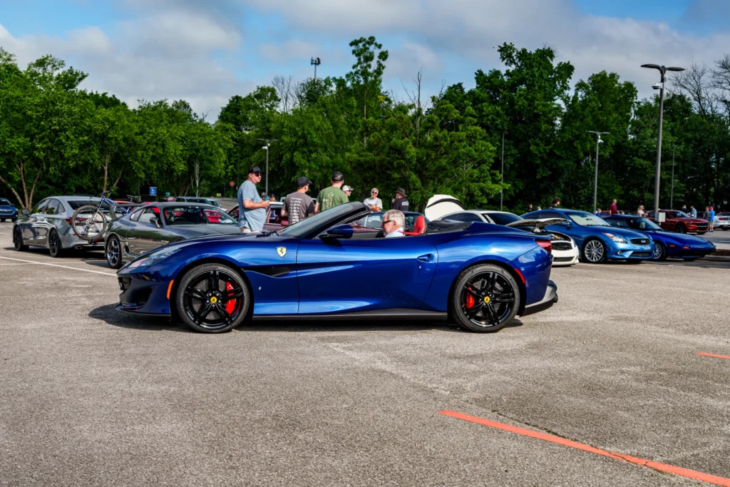 Blue Ferrari convertible with black wheels and red brake calipers parked in a lot with other cars and people
