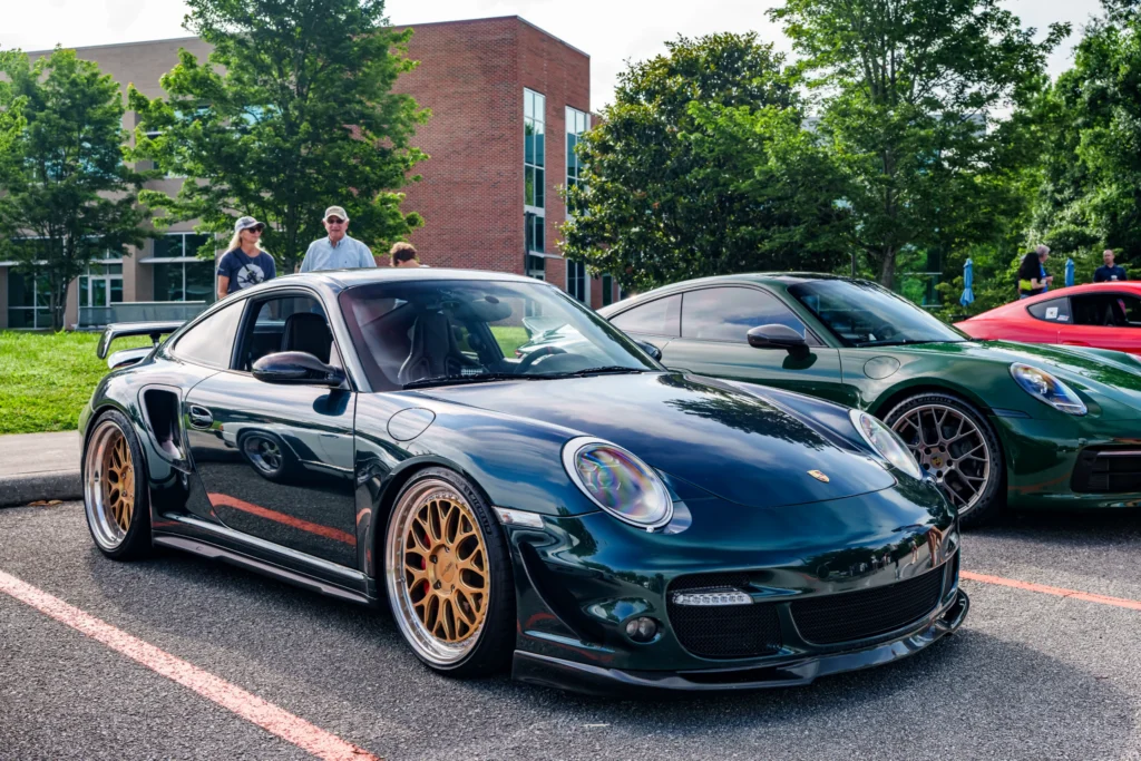 Dark green Porsche 911 with gold wheels in parking lot at a car event with trees and people in background