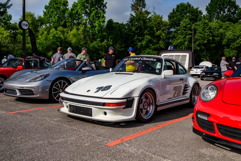 White Porsche 911 with 'Air Cooled Classics' windshield banner and several other sports cars in a parking lot at a daytime ca