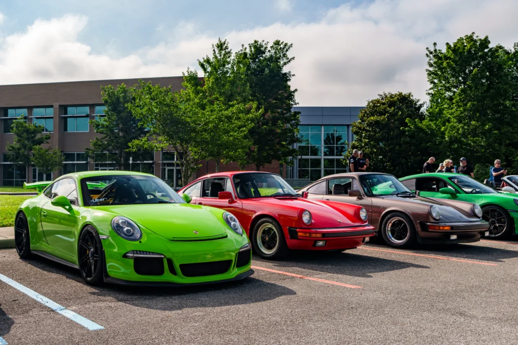 Green Porsche 911 GT3, red Porsche 911, brown Porsche 911 parked outdoors at a car event with people and trees in the backgro