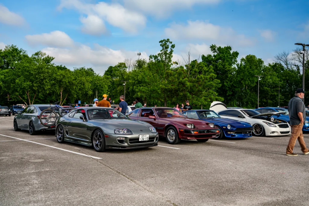 Gray Toyota Supra, maroon Mazda RX-7, and blue Datsun 240Z parked with other vehicles and people in a parking lot at a car me