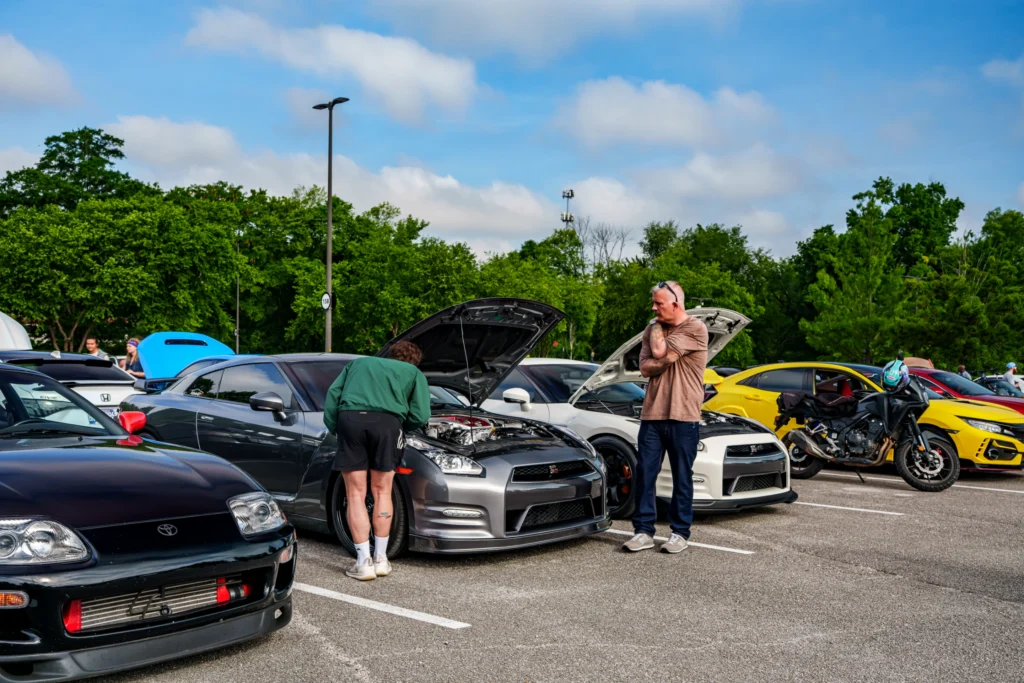 Two men inspect the engines of gray and white Nissan GT-Rs in a parking lot with other cars and motorcycles nearby under a pa