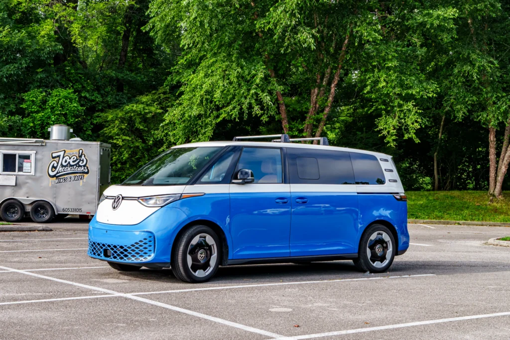 Blue and white Volkswagen electric van parked in a lot near food truck and trees