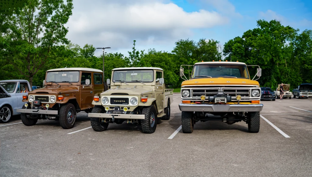 Two Toyota Land Cruisers, one brown and one beige, and a yellow Ford truck parked side by side at an outdoor car event.