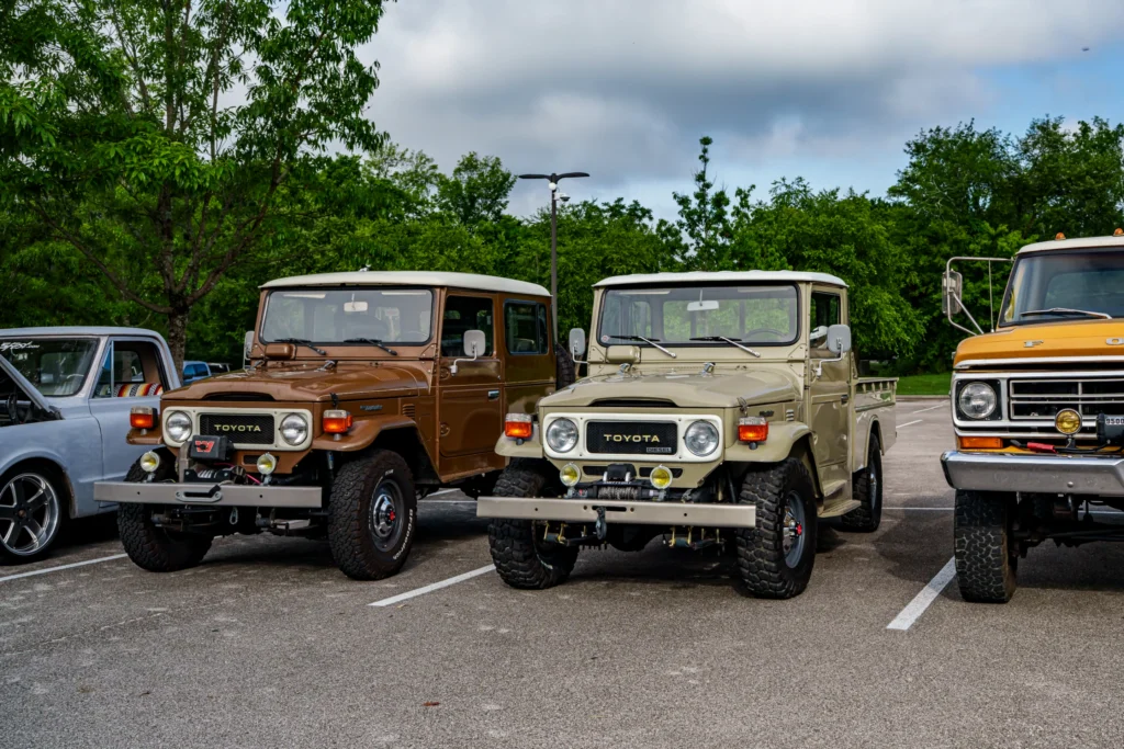 Brown and beige Toyota Land Cruisers parked side by side in a lot with trees in the background under a cloudy sky