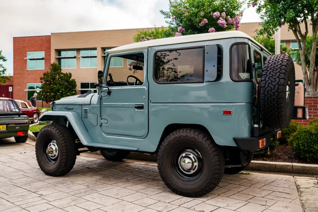 Light blue Toyota Land Cruiser parked outdoors near trees and a brick building, with a white roof and spare tire on the back