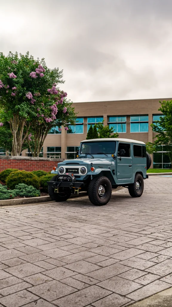Light blue Toyota Land Cruiser parked on a paved area near bushes and flowering trees with a brick building in the background