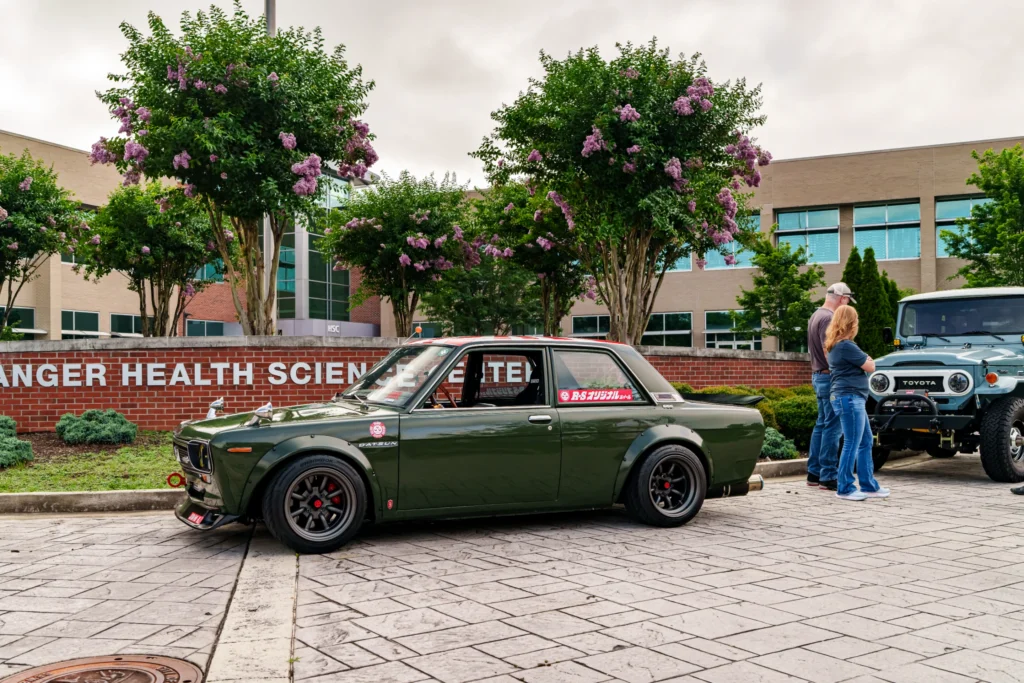 Dark green Datsun 510 parked outside a health sciences center with a blue Toyota SUV and two people nearby