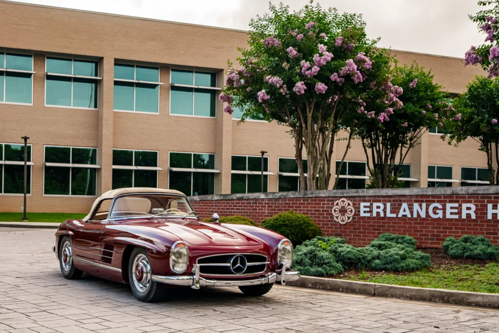 Burgundy Mercedes-Benz convertible parked in front of a brick sign with a hospital building in the background
