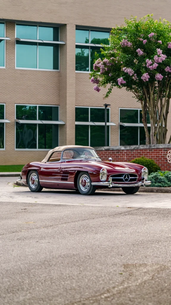 Maroon Mercedes-Benz convertible with beige soft top parked by brick wall and blooming tree in front of office building