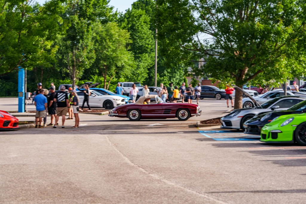 Maroon Mercedes-Benz 190 SL driving past parked cars and spectators at an outdoor car meet with trees in background