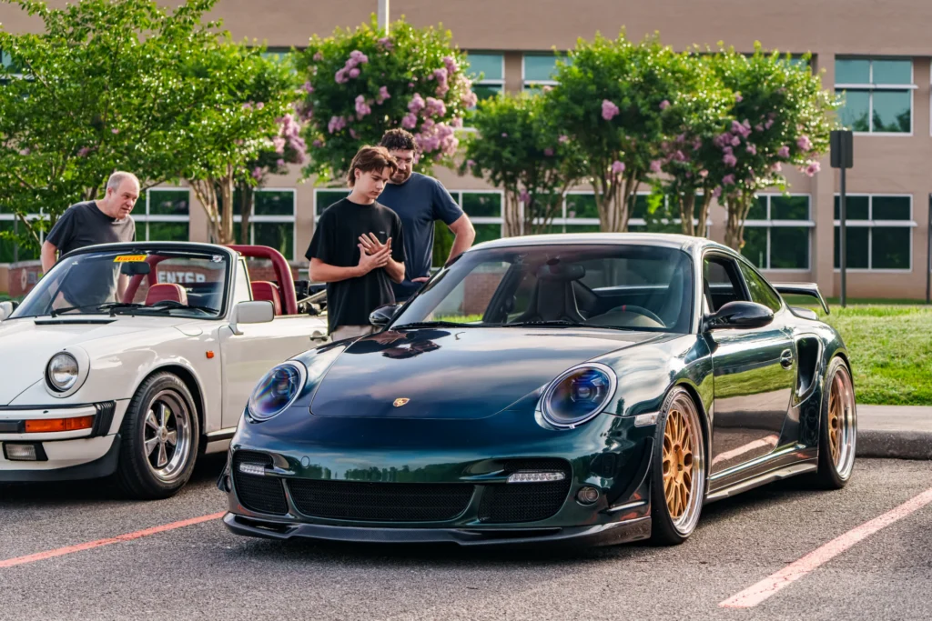 Black Porsche 911 with gold wheels parked next to a white convertible Porsche with red interior, people observing the cars in