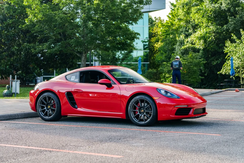 Red Porsche Cayman parked in an outdoor lot with trees and a man standing in the background