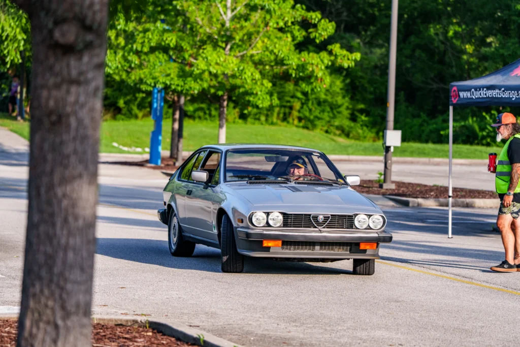 Silver Alfa Romeo GTV driving on paved road with man in safety vest holding a cup nearby