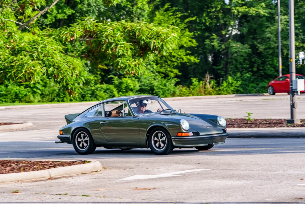 Green Porsche 911 driving in parking lot with driver wearing sunglasses on a sunny day