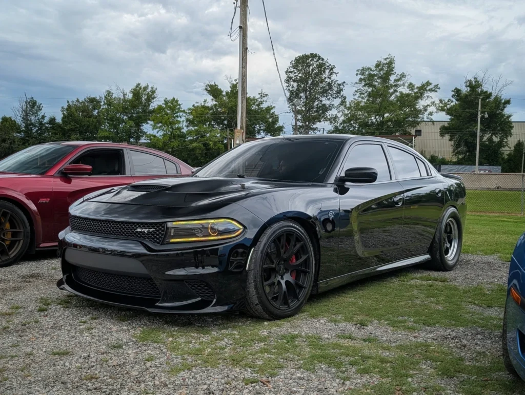 Black Dodge Charger SRT and red Dodge Charger 392 HEMI parked on gravel near trees and a utility pole under cloudy sky