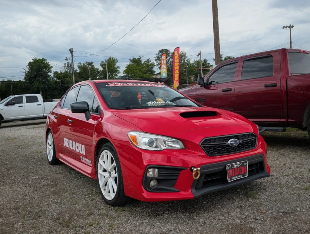 Red Subaru WRX sedan with white wheels parked on gravel near utility poles and signage for shaved ice and lemonade
