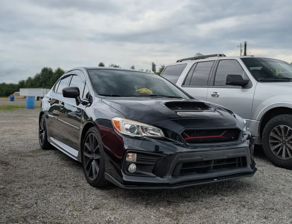 Black Subaru WRX parked next to a white SUV in an outdoor gravel lot under cloudy sky