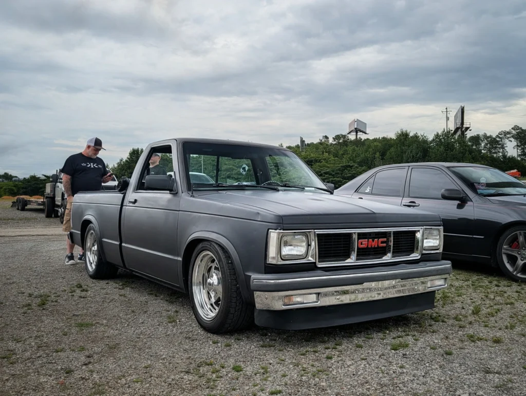 Gray GMC pickup truck parked on gravel with a man standing beside it and a black sedan nearby under cloudy sky