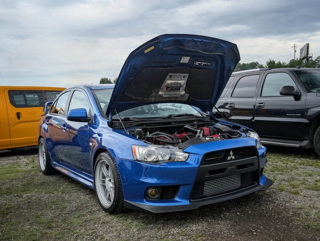 Blue Mitsubishi Lancer Evolution with hood open at a car event parking area with other vehicles in the background