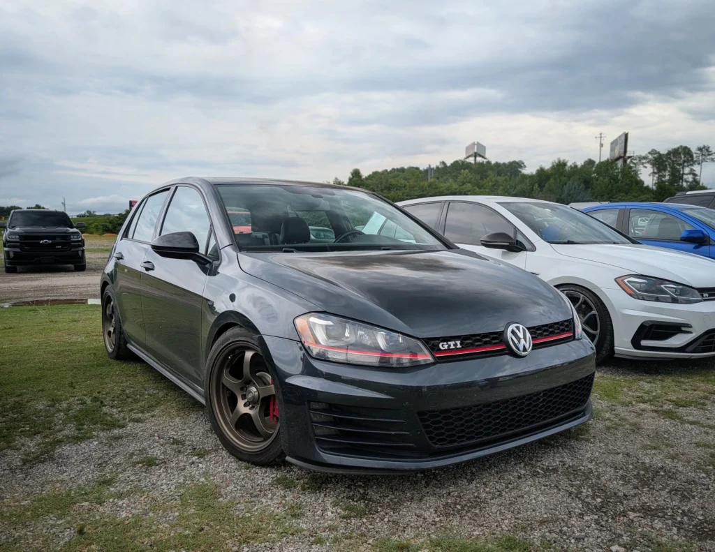 Black Volkswagen Golf GTI parked on grass with other cars and a cloudy sky background