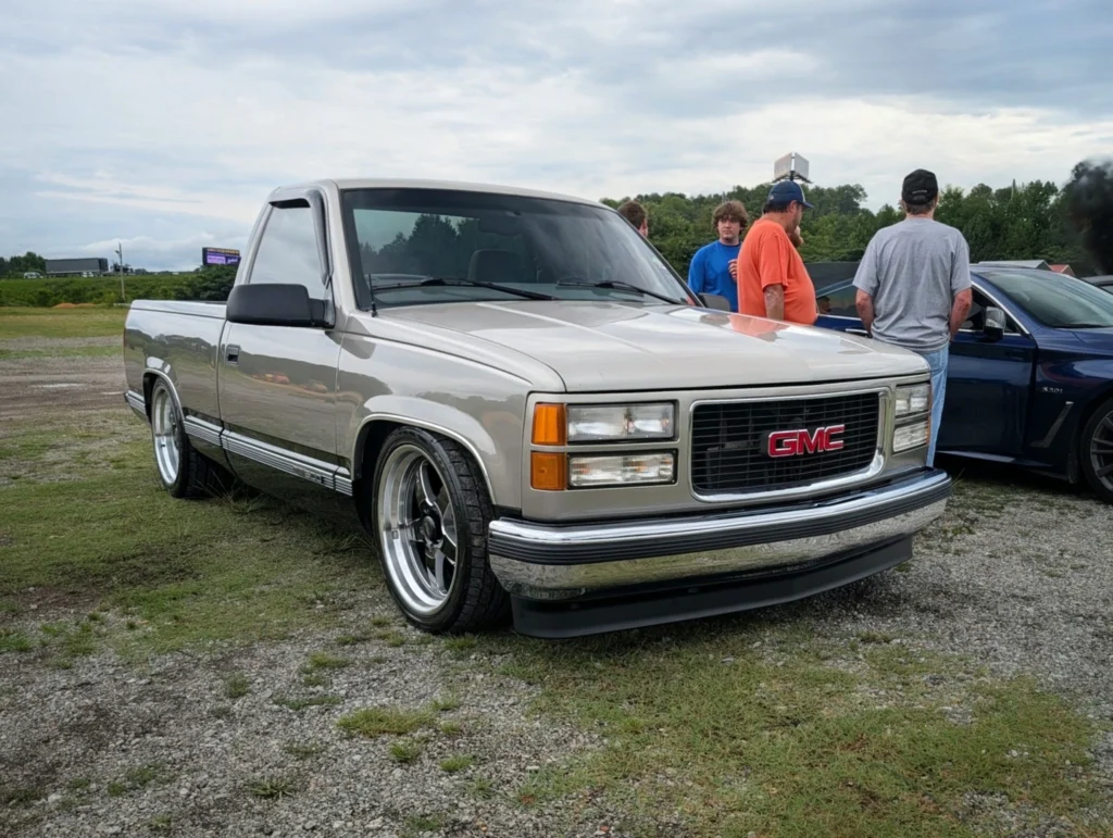 Silver GMC pickup truck parked on grass with a group of people standing nearby under a cloudy sky