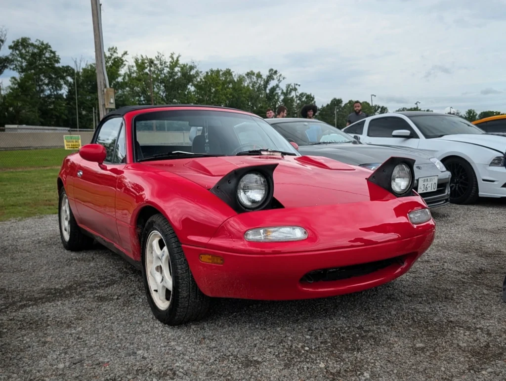 Red Mazda Miata with pop-up headlights raised in a gravel lot with other cars and people in the background