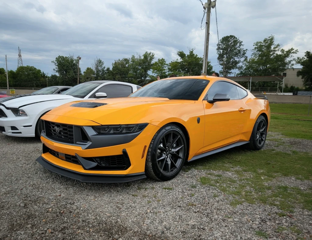 Orange Ford Mustang and white Ford Mustang parked on gravel near grassy area with trees and power lines in background